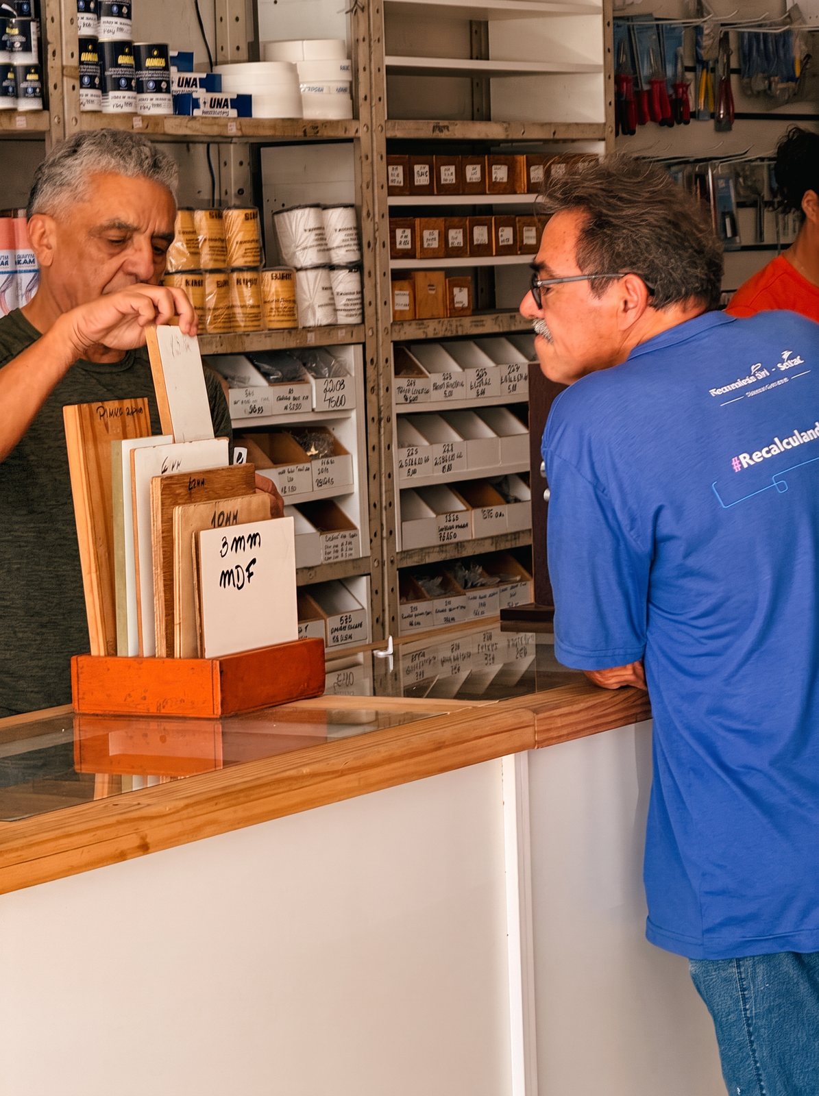 Gilmar atendendo um cliente no balcão da loja, mostrando amostras de MDF e madeira para escolher a peça certa